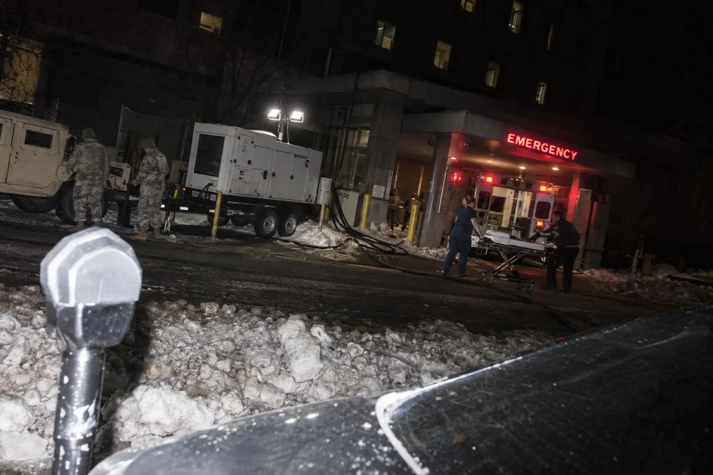 A hospital in a citywide blackout at night, running on a trailer generator, an ambulance at the emergency entrance and a National Guard humvee at the curb