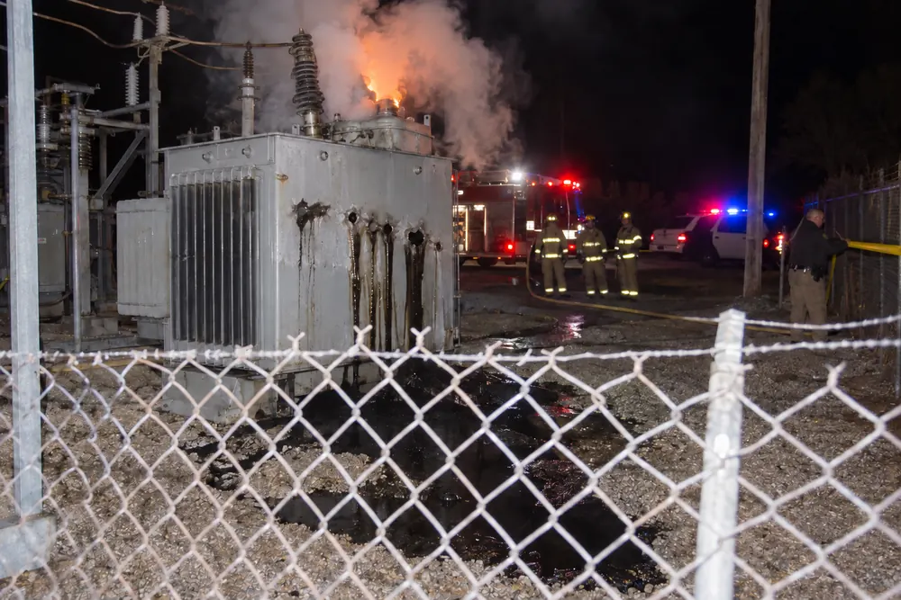 A substation transformer at night just after a rifle attack, oil pouring from its bullet holes and smoke boiling from its top, firefighters and a deputy stringing tape behind it
