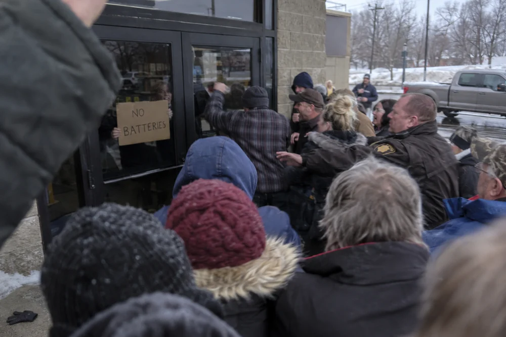 An angry crowd pounding on the locked door of a hardware store in a winter blackout, a deputy pushing them back, a worker inside holding up a cardboard sign reading No Batteries