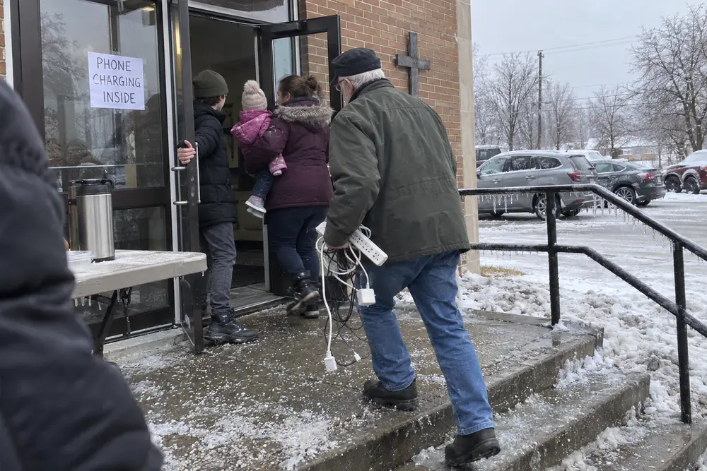 A hand-lettered poster reading PHONE CHARGING INSIDE taped inside the glass door of a church hall during an ice-storm outage, people going in and out over salted, iced steps