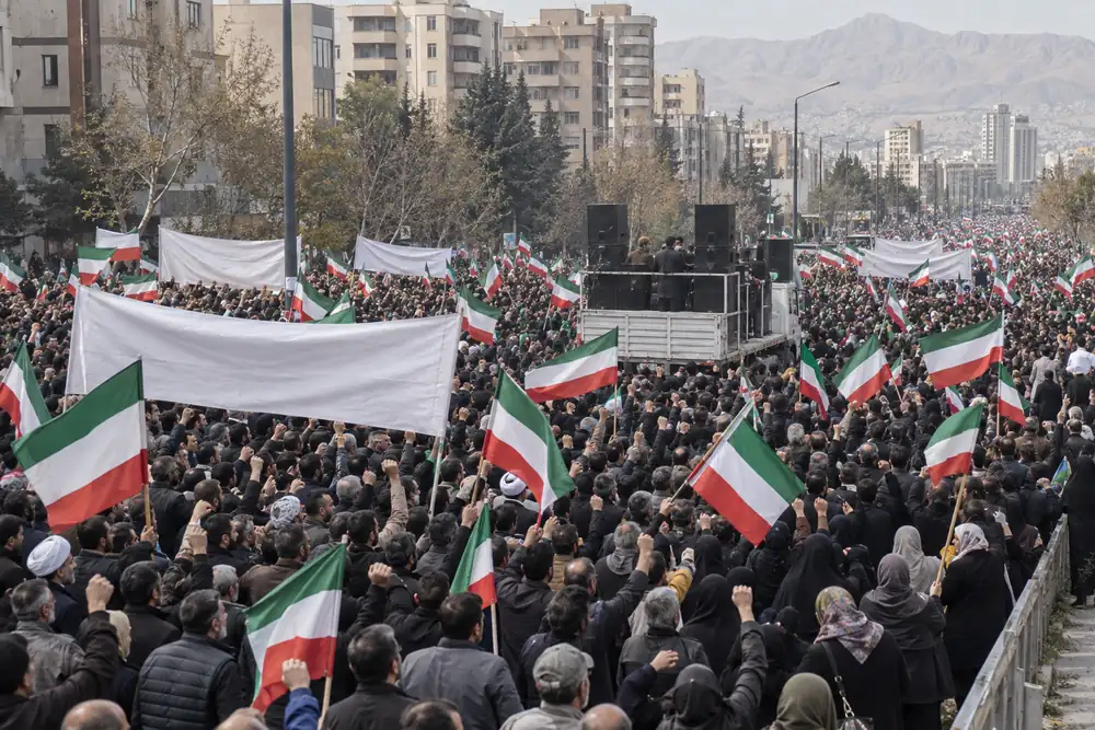 A huge daytime rally in a Middle Eastern capital: thousands of people with raised fists and plain red, white and green flags around a loudspeaker truck, apartment blocks and mountains behind