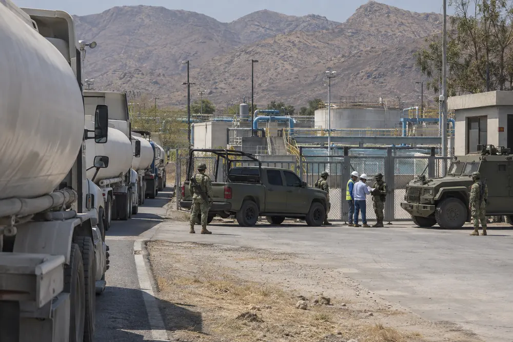 Soldiers and an armored truck guarding the gate of a big city's water plant below the mountains, water trucks lined up outside