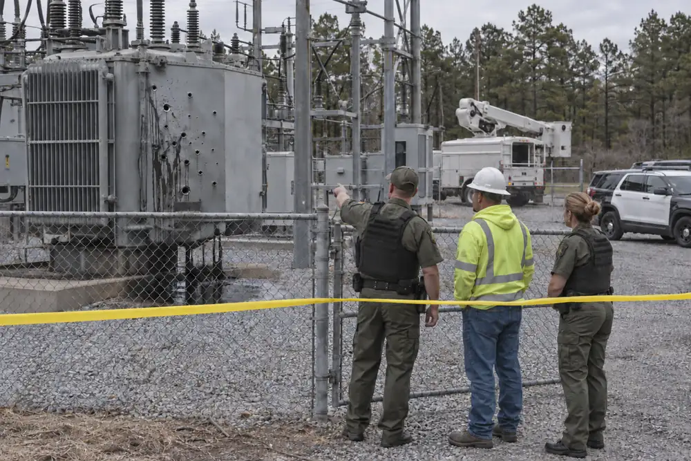 Deputies and a utility worker behind police tape at a substation, one pointing at a transformer punched with bullet holes and leaking oil