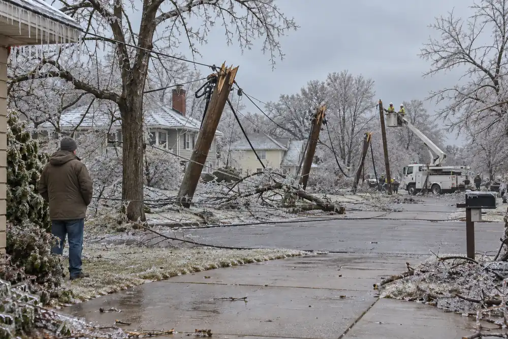 A street after an ice storm: snapped utility poles and lines down, a bucket-truck crew working and a neighbor watching