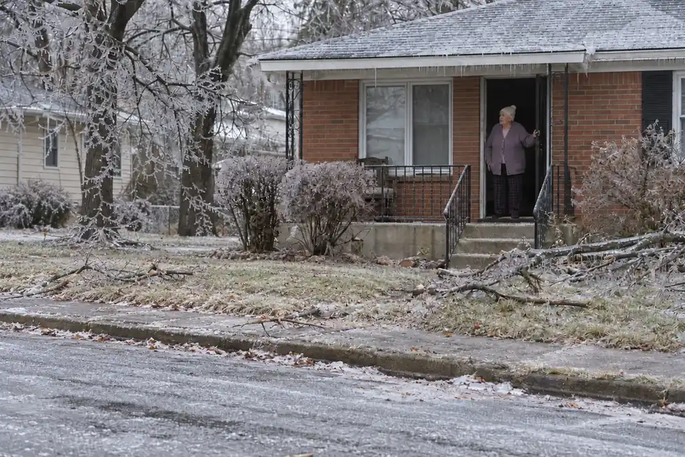 After an ice storm, an older woman alone in her dark open doorway looking out at the empty iced street, fallen branches in the yard