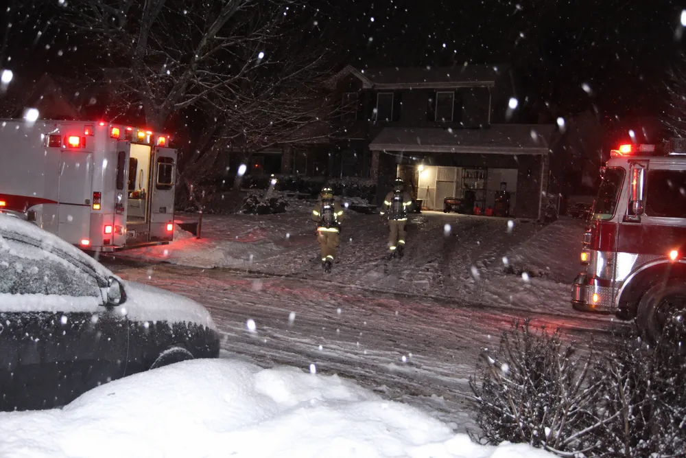 Fire crews and an ambulance at a dark snowy house at night, a portable generator and gas cans in its open garage