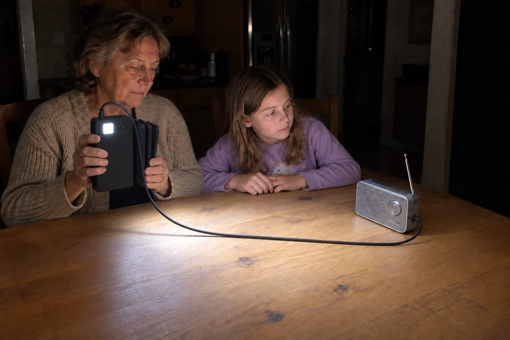 A kitchen at night with the power off: a woman holds up her folded TerraCell, the square LED light on its back lighting the table, a radio plugged into it and her granddaughter next to her