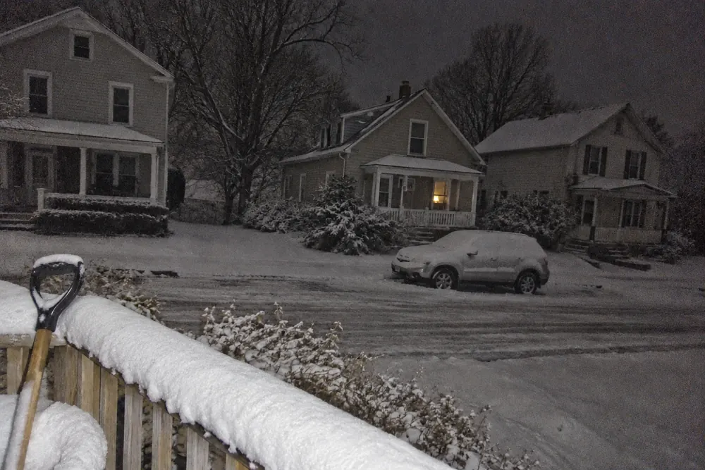 Three houses across a snowy street at night: the middle one with one small light glowing in its window, the others dark