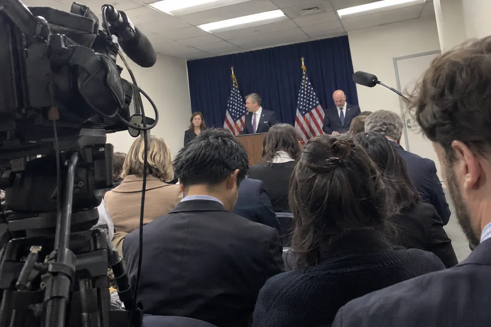 Federal officials at a podium in a crowded Washington briefing room, seen past reporters and press cameras