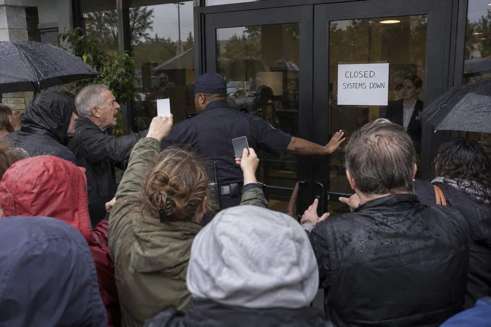 Angry customers pressing at the locked doors of a dark bank, a police officer holding them back, a handwritten sign reading Closed, Systems Down