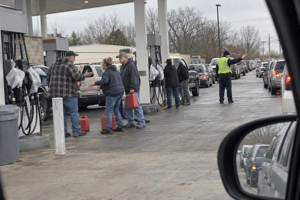 A gas station during the shortage: a long line of cars, people with gas cans arguing at a pump and a police officer directing traffic