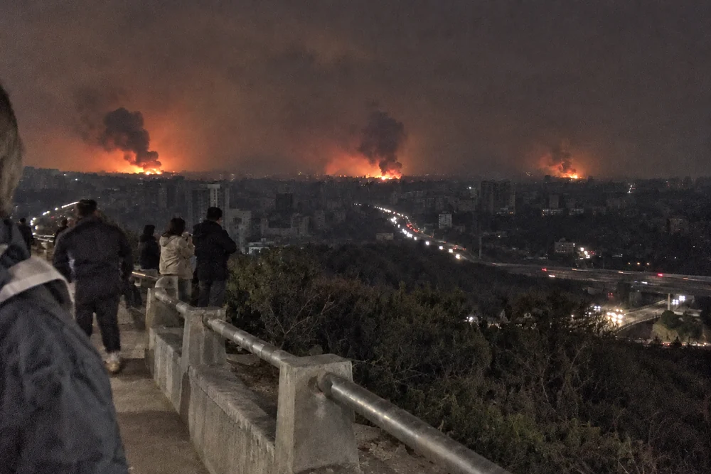 A city at night seen from a hill, every window and streetlight dark, lit only by three fires and the headlights on its highways