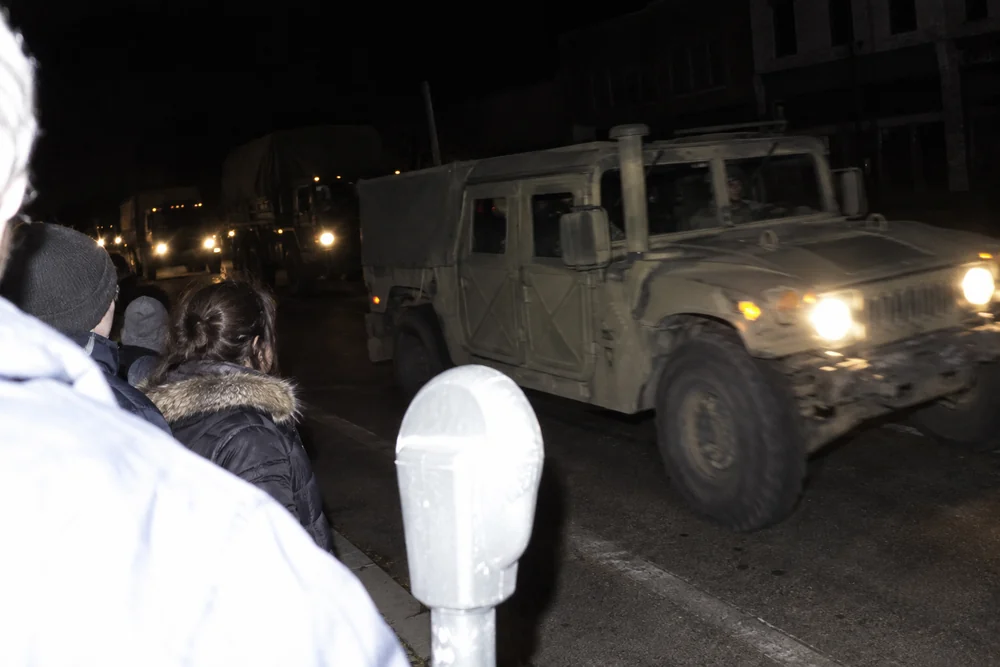 National Guard trucks rolling down the dark main street of a small town in a blackout, residents watching from the sidewalk