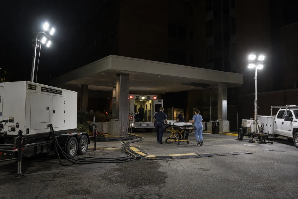 A hospital emergency entrance at night lit only by portable floodlight towers, a trailer generator and cables, an ambulance backed in