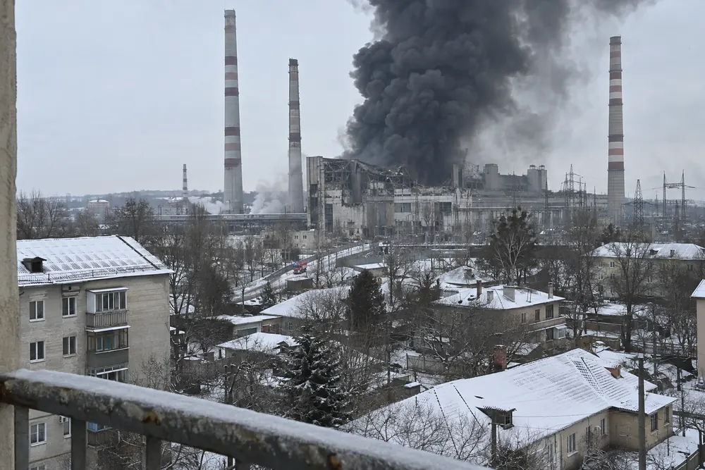 A city power plant hit and smoking, a column of black smoke over the snowy rooftops, seen from an apartment balcony