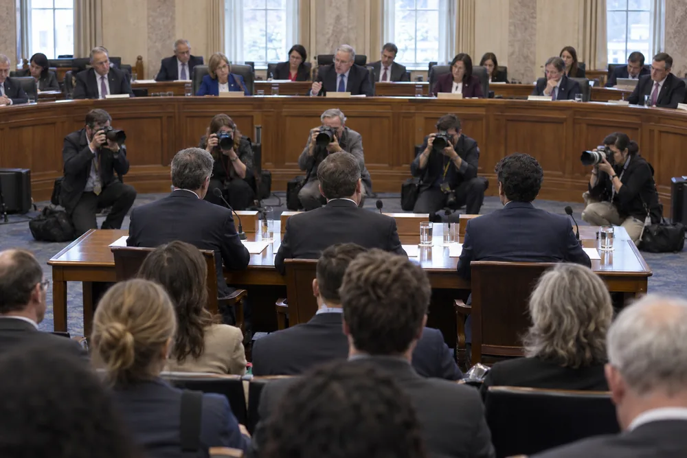 A congressional hearing seen from the back of the room: three witnesses at the table, lawmakers on the dais, news photographers kneeling between them