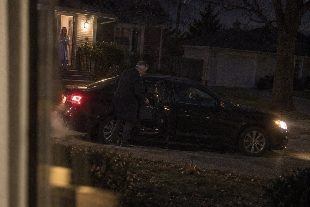 Before sunup, seen through a neighbor's front window: Curtis in a dark overcoat putting a bag into the back seat of a black car idling at the curb, his wife in a robe watching from the lit doorway