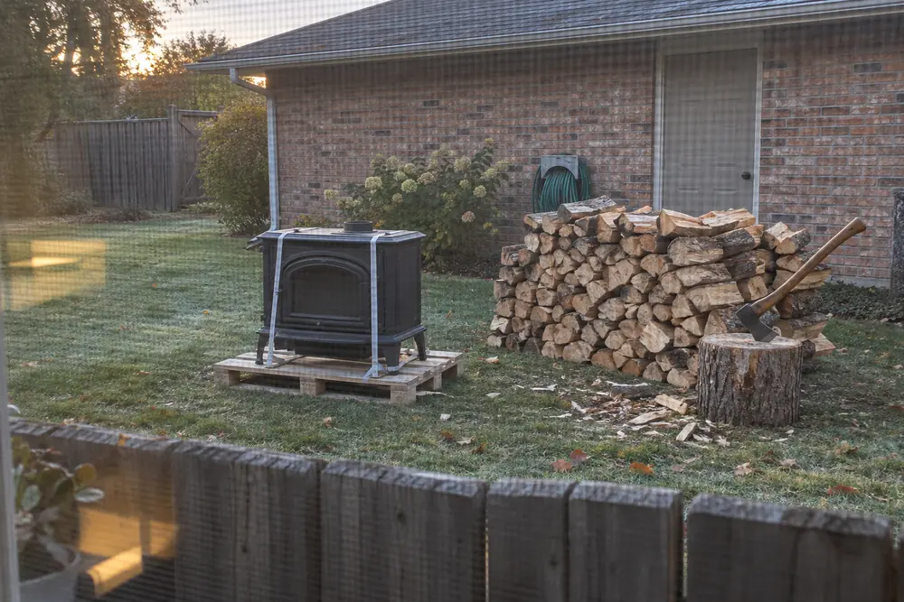 Through a kitchen window screen at dawn: a new wood stove still strapped to a pallet in the neighbor's side yard, a pile of split oak and an axe in a chopping stump