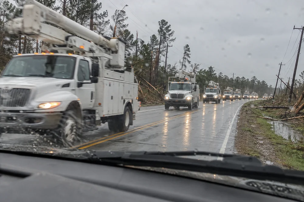 A long convoy of white utility bucket trucks rolling down a highway into a storm-hit town on a grey morning, snapped trees on the shoulder