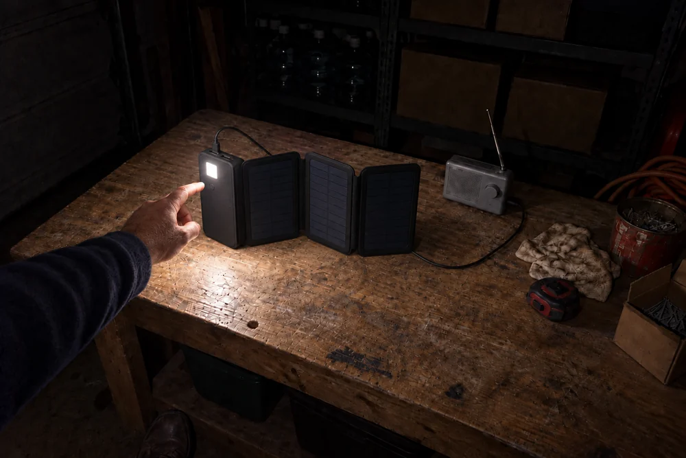 A dark garage workbench lit only by the square LED light on the back of a TerraCell standing open on the bench, a man's finger just leaving its power button, a radio cabled to it