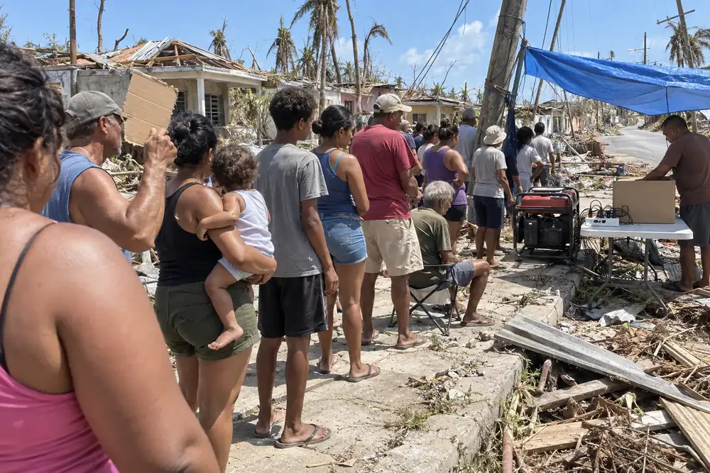 Weeks after a hurricane on a tropical island: a long line of people in the hot sun waiting their turn at a gas generator under a blue tarp, a power strip of charging cables on a folding table, snapped poles and torn roofs along the street