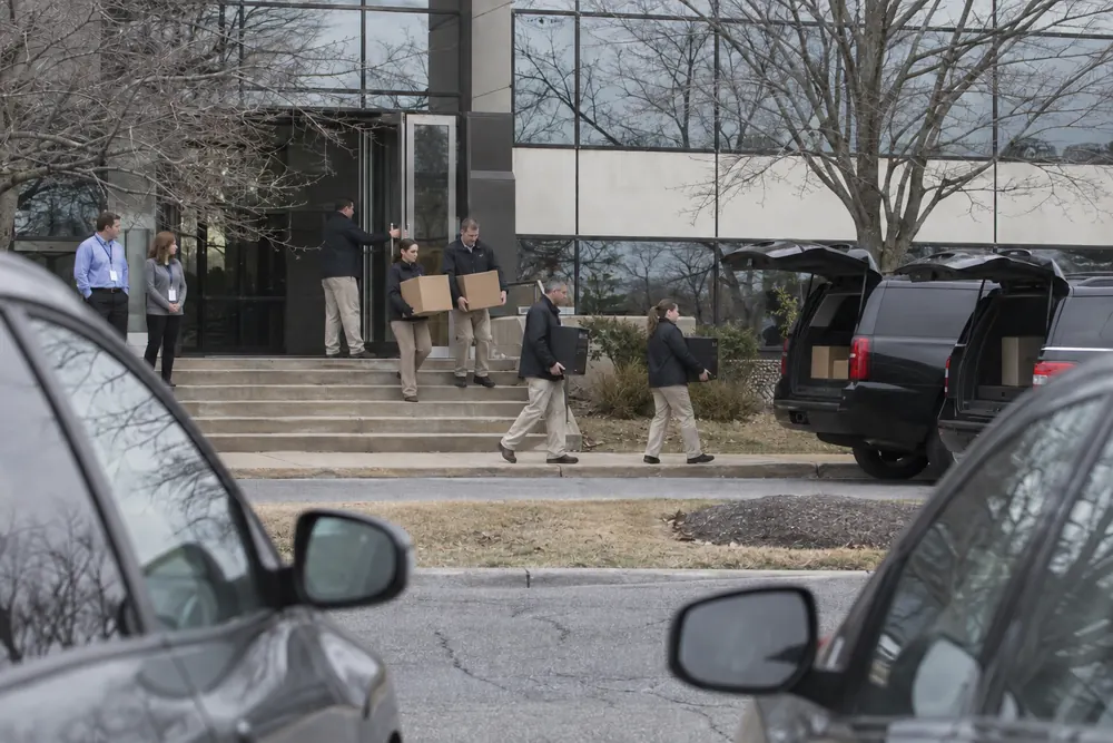 Federal agents carrying evidence boxes and computer towers down the steps of an office building to dark SUVs, two office workers watching, seen from across the street