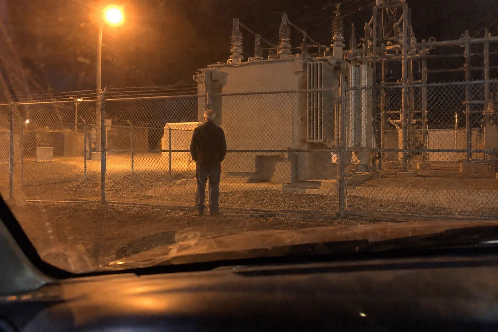 A photo through a pickup windshield at night: a tall older man alone at a substation fence, facing the giant transformer under an orange light
