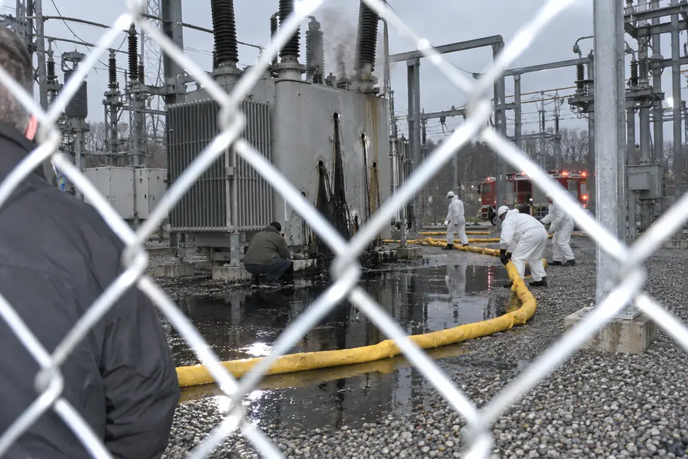 A substation at first light after a rifle attack: oil running from bullet holes in a giant transformer into a black lake on the gravel, a hazmat crew laying yellow booms, one transformer smoking