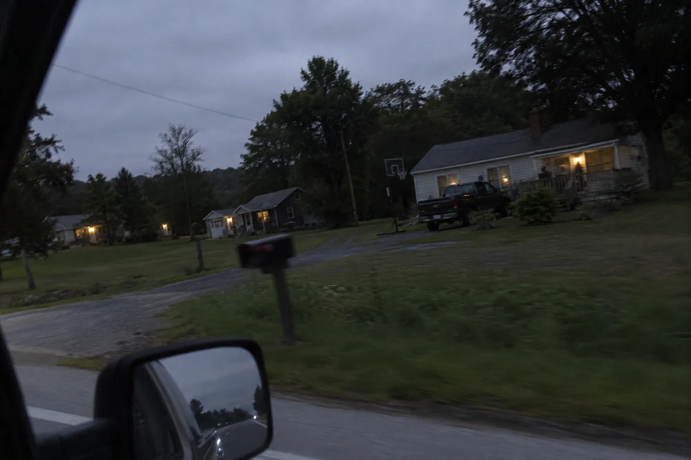 A rural road at dusk seen from a truck window, every porch light along it burning