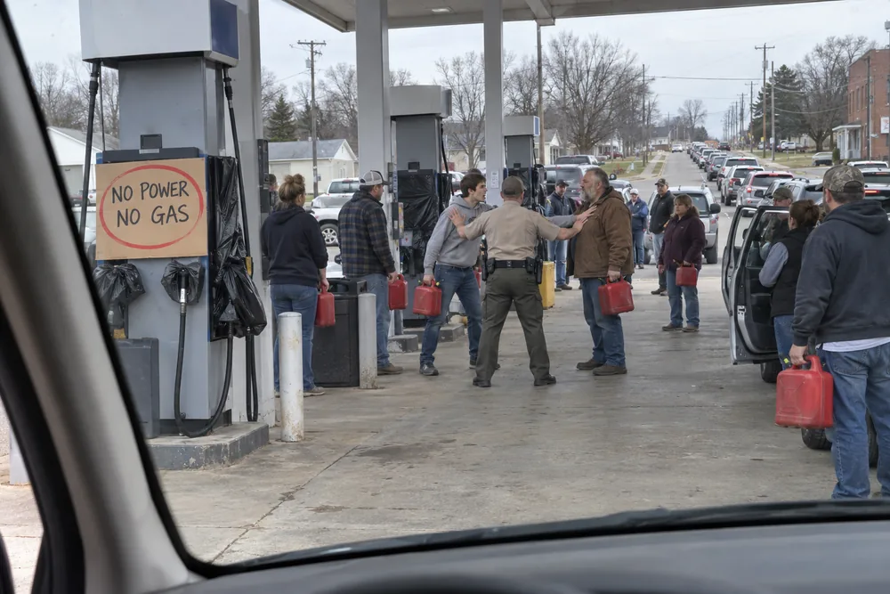 Through a windshield at a dead gas station in a blackout: a sign reading No Power No Gas circled in red, two men with empty gas cans shoving each other as a deputy pushes between them, a line of cars down the road
