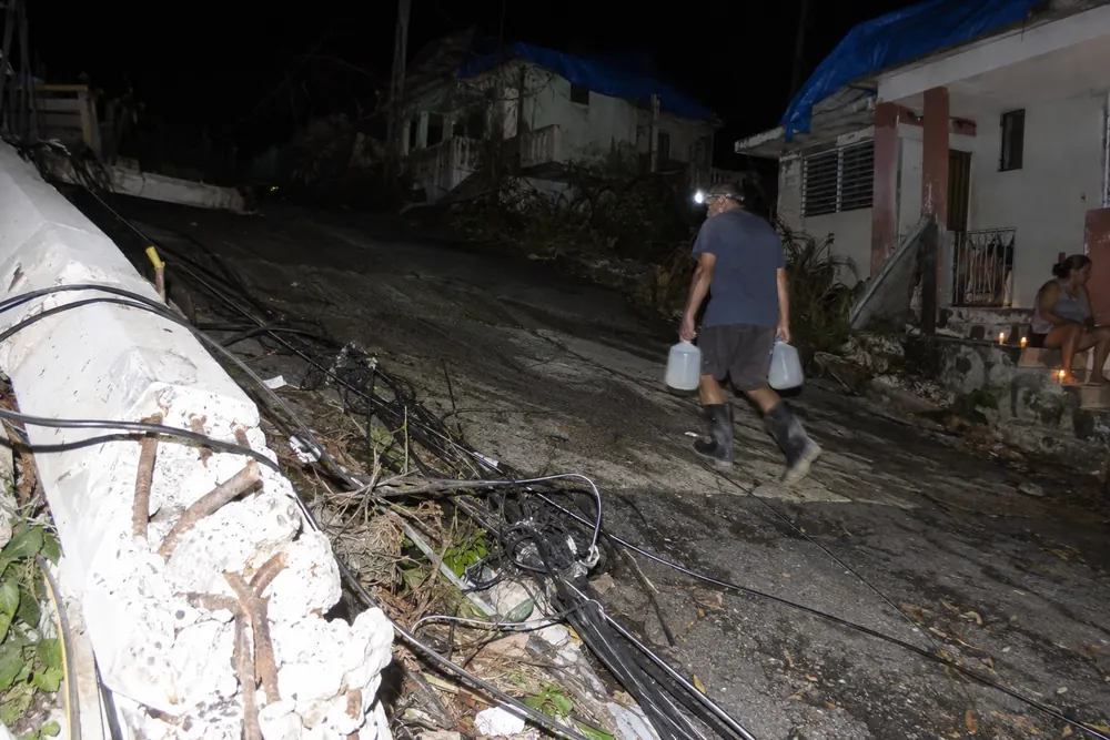 A hillside street in Puerto Rico at night months after Hurricane Maria: snapped concrete poles and tangled lines in the street, candles on a doorstep, a man carrying water jugs by headlamp