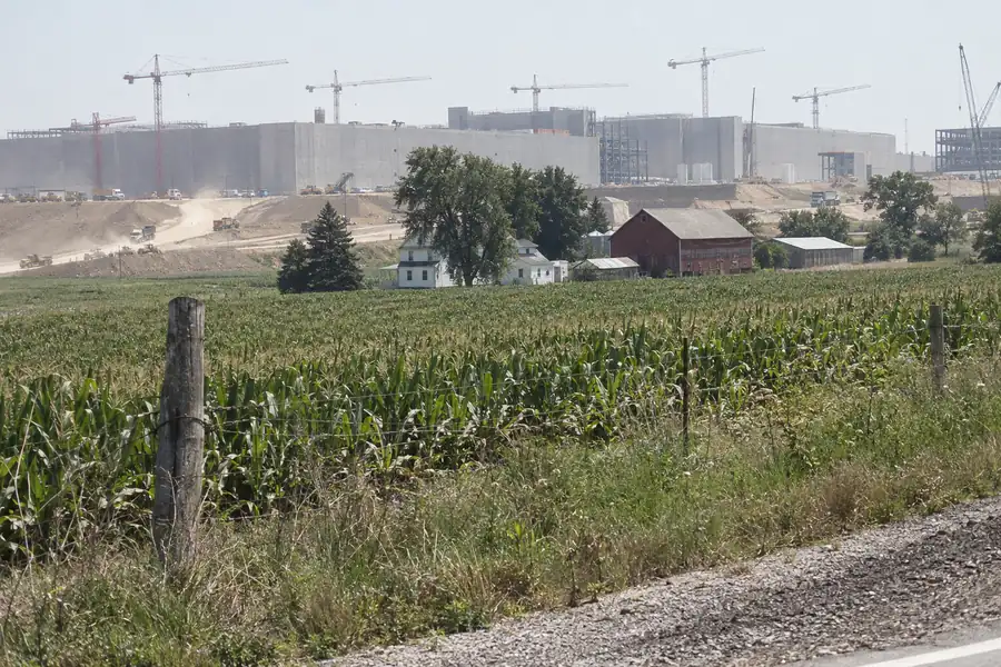A giant computer campus under construction behind a farmhouse, a red barn and a cornfield, cranes along the skyline