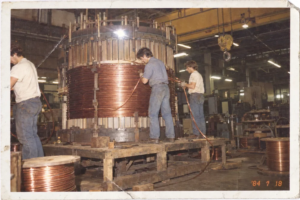A faded 1984 snapshot print: three young men winding copper wire around the core of a giant transformer on a factory floor