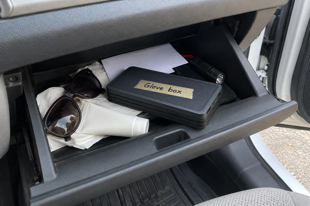 An open car glove box in daylight with napkins and sunglasses, a folded TerraCell on top with a strip of tape reading Glove box