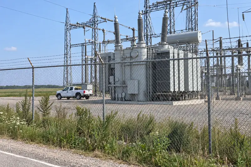 A county substation from the road in daylight, through the fence: a giant transformer two storeys tall, a white pickup tiny beside it