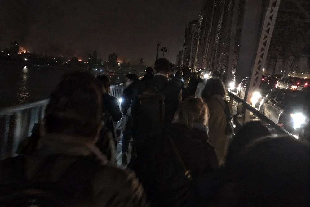 A big city in a total blackout at night, every tower dark, fires burning along the waterfront, crowds walking home across a bridge beside stalled traffic