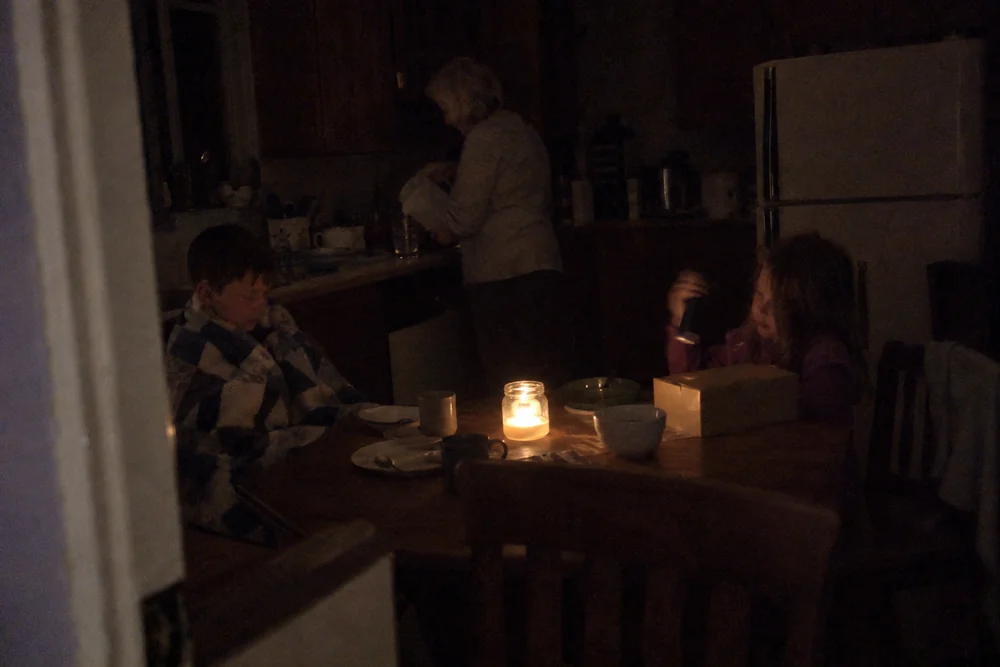A kitchen in a blackout at night: two children and their grandmother, one candle the only light, a girl shaking a dead flashlight