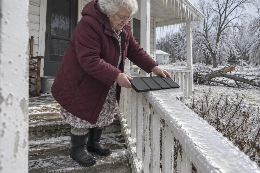 An old woman in a winter coat on her iced front porch laying an open TerraCell along the porch rail, icicles overhead and a broken limb on the lawn