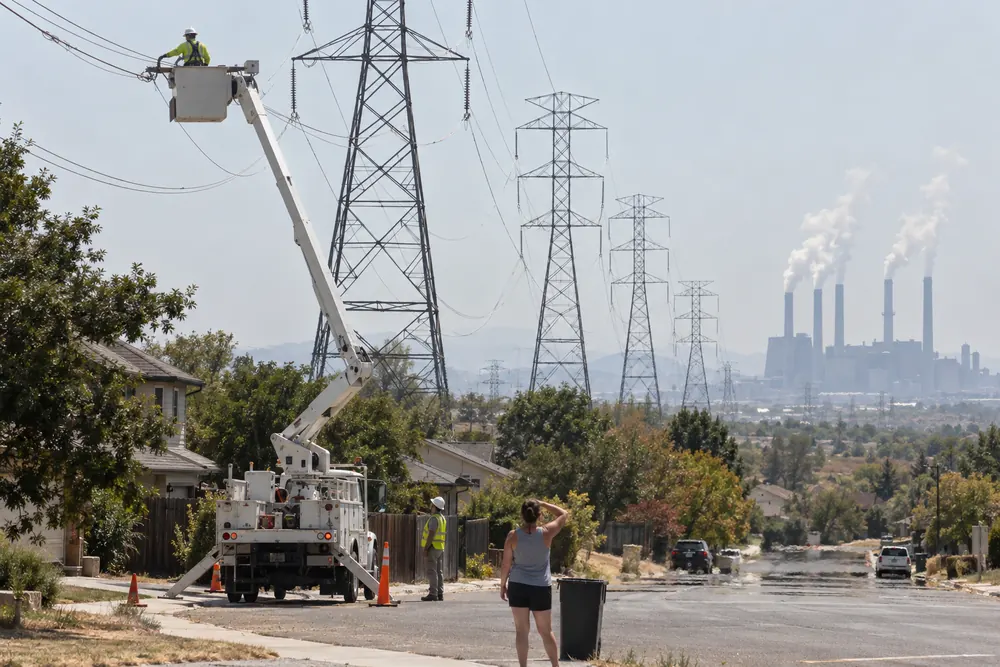A line crew's bucket truck under giant power line towers that run over a neighborhood in a heat wave, a power plant steaming in the distance, a woman shading her eyes to watch