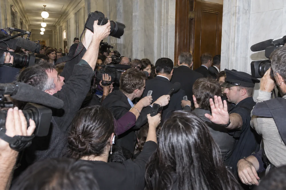 Reporters and cameras crowding a Senate office hallway as people in suits leave a closed-door briefing