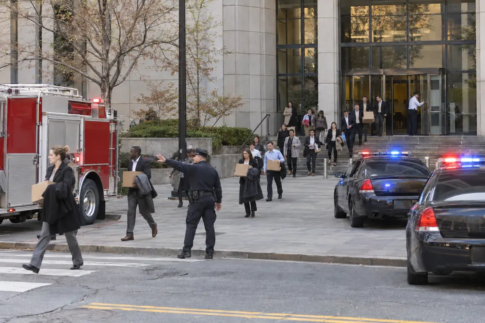 Staff evacuating a government office building in daylight, police cruisers with lights flashing and a fire truck on the plaza