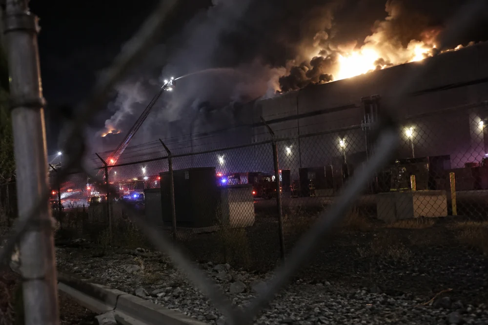 A giant data center on fire at night, flames through the roof, fire engines and a ladder truck spraying water, seen through the perimeter fence