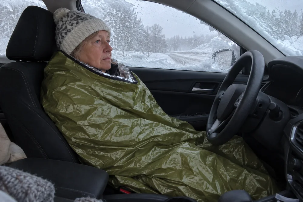 A woman in the driver's seat of a car stopped on a snowy road at dusk, inside her olive TerraShell pulled up to her shoulders, snow on the windshield