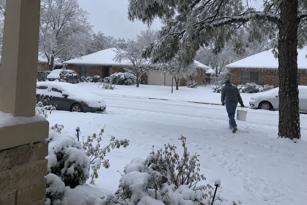 A Texas suburban street under snow the morning after the freeze: dark brick houses, icicles on the gutters, cars under snow, a man carrying a bucket of snow