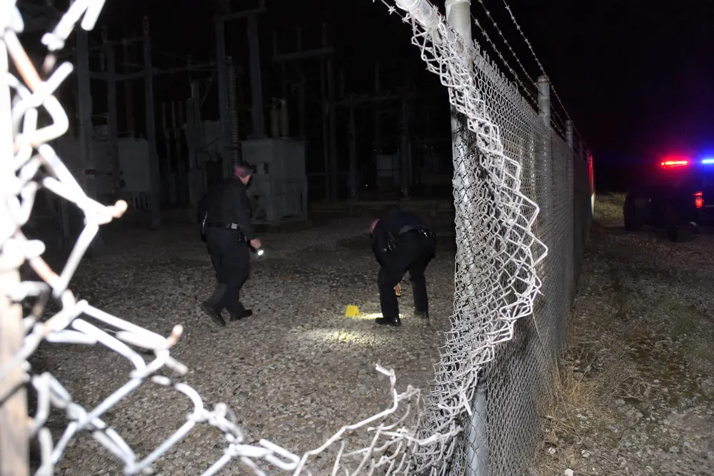 Deputies with flashlights at a hole cut in a substation fence at night, an evidence marker on the gravel