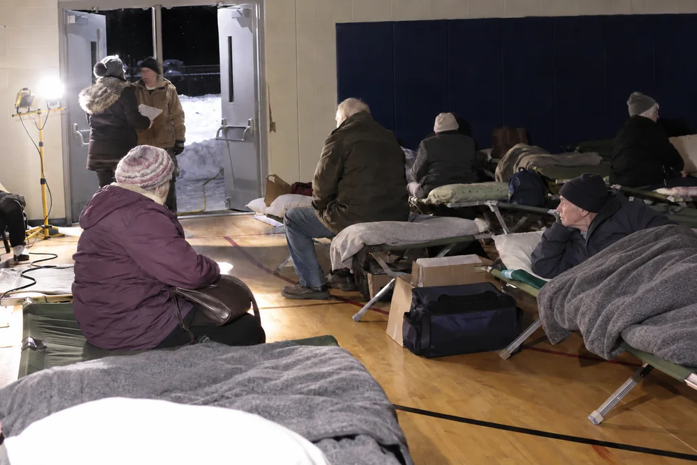 A small-town school gym used as a warming center at night: rows of cots with grey blankets, older people in coats sitting on them, a volunteer with a clipboard at the door, snow outside