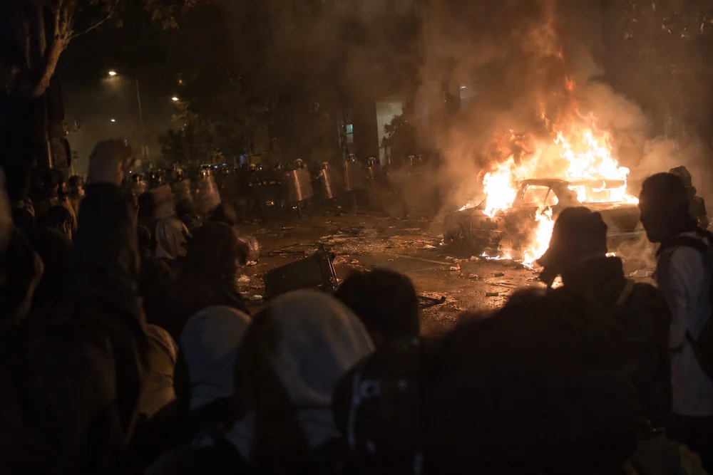 A police riot line with shields across a downtown street at night, a car burning in the road in front of an angry crowd