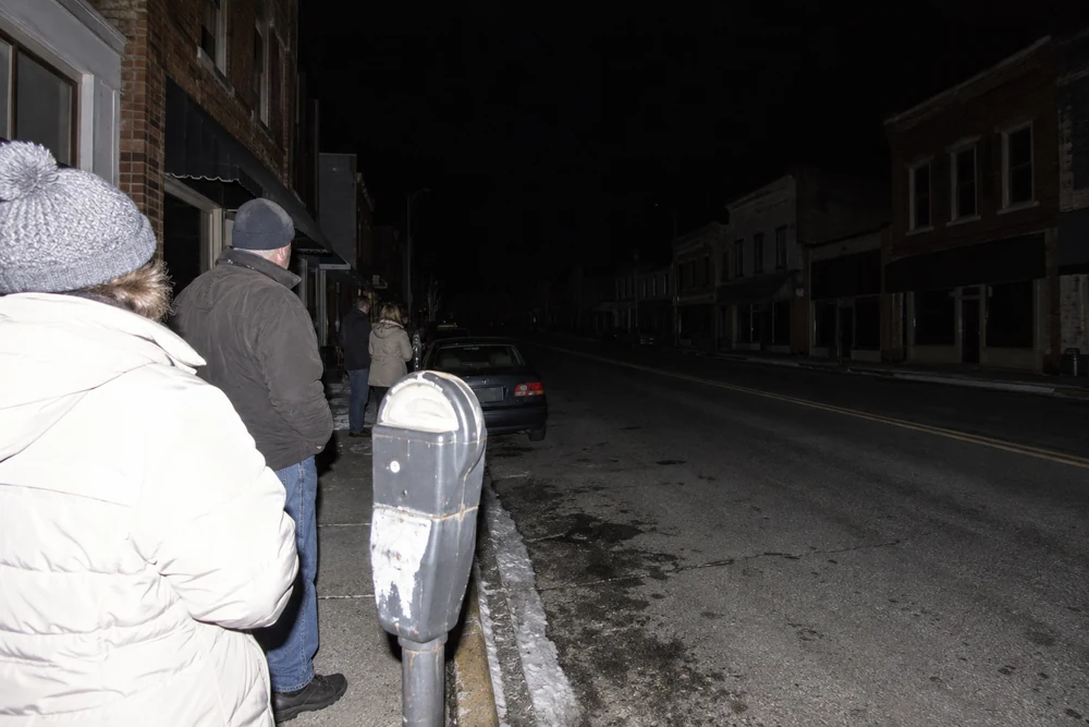 A small town's main street in a blackout at night, every shop and streetlight dark, residents in winter coats on the sidewalk looking down the empty street