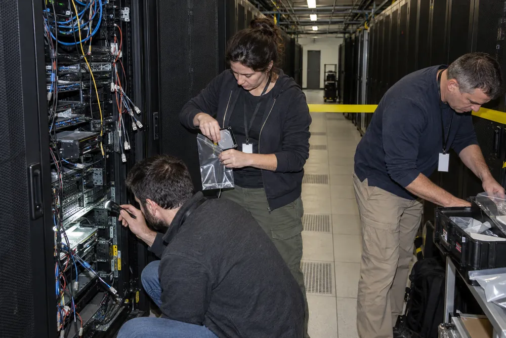 Incident responders at an open server rack under flash, cables pulled, a drive going into an evidence bag, caution tape across the aisle