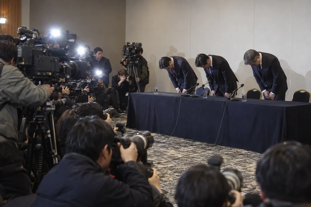 Bank executives in dark suits bowing deeply in apology at a press conference, photographers' flashes firing